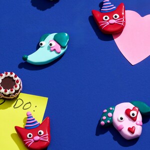Puede incluir: Una colección de imanes coloridos y caprichosos sobre un fondo azul. Los imanes incluyen un gato rojo con gorro de fiesta, un perro azul claro, un corazón rosa y pasteles en miniatura. Una nota adhesiva amarilla dice "do:"