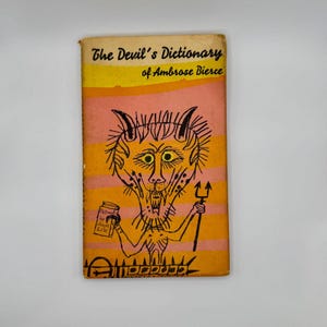 May include: The Devil's Dictionary by Ambrose Bierce, a vintage paperback book with a cartoon illustration of a devil holding a pitchfork and a can of "Pictograms by Joseph Low" on the cover.