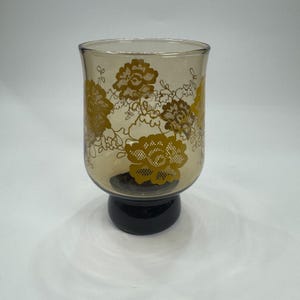 May include: A clear glass candle holder with a gold floral design. The candle holder has a black base and a wide, cylindrical shape. The floral pattern is a repeating design of large flowers and leaves.