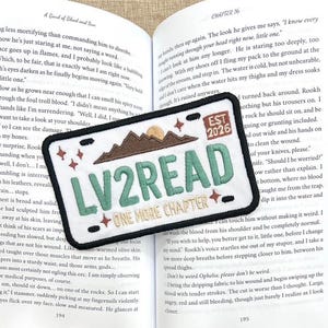 May include: A rectangular embroidered patch with a white background and black border. It features the text 'LV2READ' in large green letters, 'EST 2026' in a brown box, and 'ONE MORE CHAPTER' in gold. A brown mountain range, a gold sun, and small red stars are included.