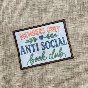 May include: A rectangular embroidered patch with a white background and a dark brown border. The text reads MEMBERS ONLY in orange, ANTI SOCIAL in blue, and book club in green, accented by two green leafy sprigs and a small blue heart.