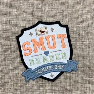 May include: A shield-shaped embroidered patch with a white background and black border. It features an open book with a red chili pepper, the orange text SMUT, a small blue heart, the green text READER, and a blue banner reading MEMBERS ONLY.