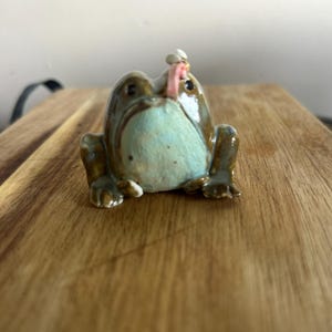 May include: A small ceramic frog figurine with a glossy brown and light blue finish. The frog is depicted with a small pink tongue extended upwards, catching a tiny white fly perched near its head. The sculpture features dark eyes and limbs.