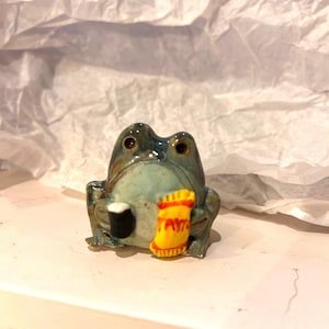 May include: A small ceramic frog figurine with a glossy blue-green glaze. The frog is sitting upright and holding a black mug in one hand and a yellow bag of crisps with the word TAYTO printed in red text in the other hand.