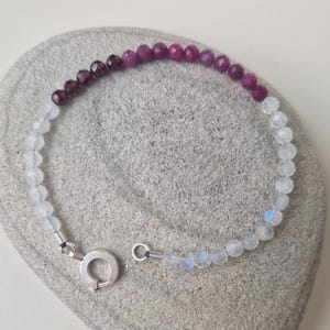 May include: A bracelet made with white moonstone and purple and red gemstone beads. The bracelet has a silver clasp.