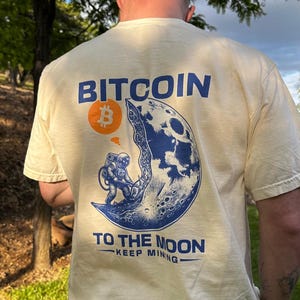 May include: Cream-colored t-shirt with a blue and orange graphic featuring the word "BITCOIN" and an astronaut on the moon. The phrase "TO THE MOON - KEEP MINING" is printed below the graphic.
