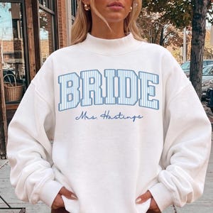 May include: A white long-sleeve sweatshirt with a mock neck collar. The front features the word BRIDE in large, blue-striped block letters, with the text Mrs. Hastings written underneath in a smaller, blue cursive script font.