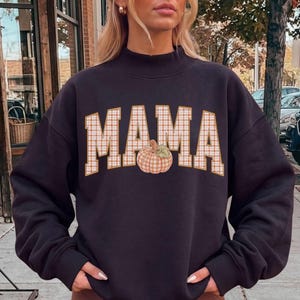 May include: A dark charcoal gray long-sleeve sweatshirt with a mock neck collar. The front features the word MAMA in large, block-style letters with a white and orange plaid pattern, and a small illustration of a pumpkin replacing the letter A.
