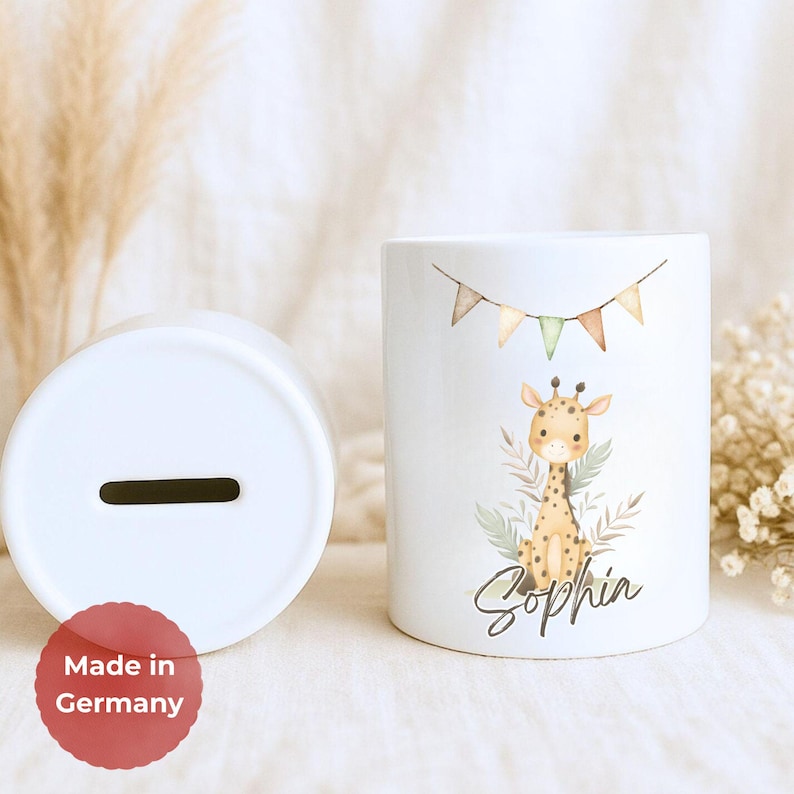 May include: White ceramic money box with a slot on top and a giraffe illustration. The giraffe is surrounded by greenery and a banner with colourful flags. The name "Sophia" is written below the giraffe. Made in Germany.