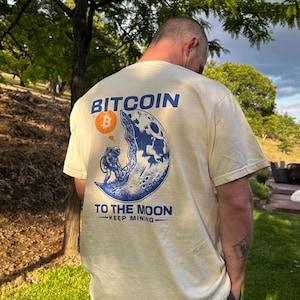 May include: Cream-colored t-shirt with a Bitcoin graphic on the back. The design features a blue and white illustration of an astronaut on the moon, with the words "Bitcoin" and "To The Moon" printed above and below the image.