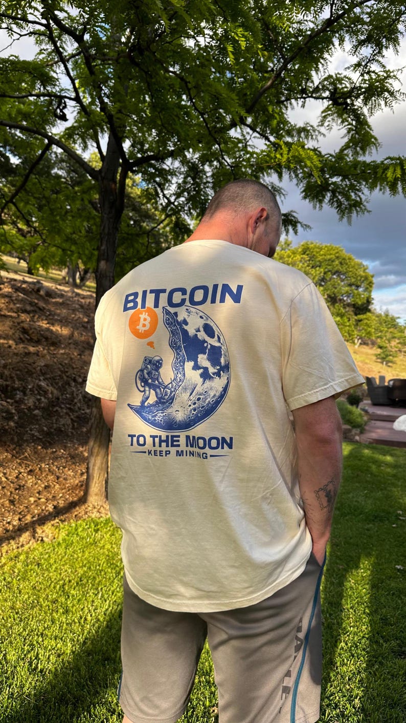 May include: Cream-colored t-shirt with a Bitcoin graphic on the back. The design features a blue and white illustration of an astronaut on the moon, with the words "Bitcoin" and "To The Moon" printed above and below the image.