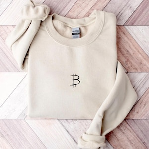 May include: A cream-colored sweatshirt with a black Bitcoin symbol embroidered on the front. The long sleeves are folded, and the garment is made of a soft material. The sweatshirt is laid on a wooden surface.