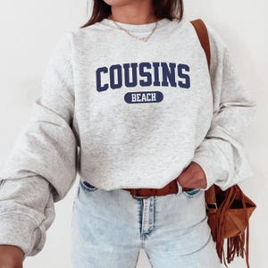 May include: A light grey sweatshirt featuring the navy blue text "COUSINS BEACH". The sweatshirt has long sleeves and a crew neck. The model is wearing light blue jeans and a brown belt, with a brown bag.