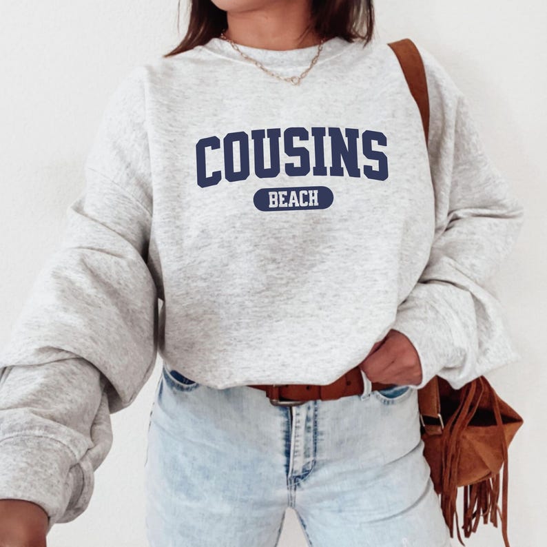 May include: A light grey sweatshirt featuring the navy blue text "COUSINS BEACH". The sweatshirt has long sleeves and a crew neck. The model is wearing light blue jeans and a brown belt, with a brown bag.