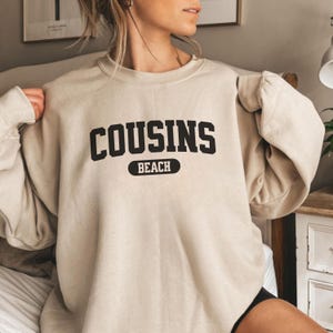 May include: A beige sweatshirt featuring the text "COUSINS BEACH" in black. "BEACH" is in a black oval below "COUSINS". The long-sleeved top is a neutral colour, suitable for casual wear.