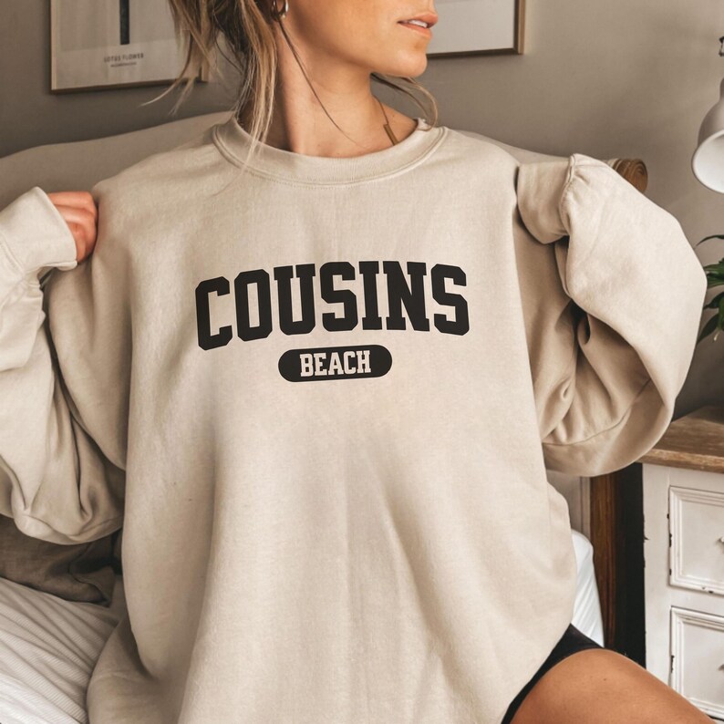 May include: A beige sweatshirt featuring the text "COUSINS BEACH" in black. "BEACH" is in a black oval below "COUSINS". The long-sleeved top is a neutral colour, suitable for casual wear.