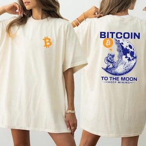 May include: Cream-colored t-shirt with a Bitcoin logo on the front and a graphic on the back. The back graphic features an astronaut on the moon with the words "Bitcoin To The Moon" and "Keep Mining".