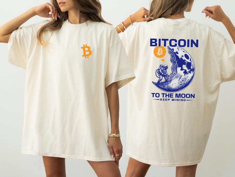 May include: Cream-colored t-shirt with a Bitcoin logo on the front and a graphic on the back. The back graphic features an astronaut on the moon with the words "Bitcoin To The Moon" and "Keep Mining".