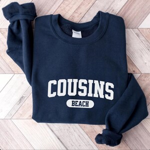 May include: A navy blue sweatshirt featuring the text "COUSINS BEACH" in white, block lettering. The garment is folded, displaying the cuffs and neckline. The text is centrally positioned on the chest.