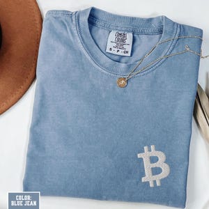 May include: A blue jean colored t-shirt with a white embroidered Bitcoin symbol. The shirt has a crew neck and short sleeves. A gold necklace with a pendant is draped across the shirt. The tag reads "Comfort Colors".