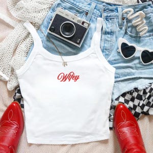 May include: A white tank top with the word Wifey embroidered in red script, paired with light blue denim shorts, red leather cowboy boots, a black and silver film camera, white heart-shaped sunglasses, and a white crochet cardigan.