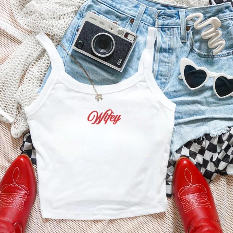 May include: A white tank top with the word Wifey embroidered in red script, paired with light blue denim shorts, red leather cowboy boots, a black and silver film camera, white heart-shaped sunglasses, and a white crochet cardigan.