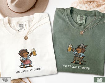May include: Two short-sleeve t-shirts in ivory and moss green. Each shirt features a graphic of a dachshund holding a beer mug with the text WE PROST AT DAWN. The ivory shirt shows a dachshund in a dirndl, while the moss shirt shows one in lederhosen.