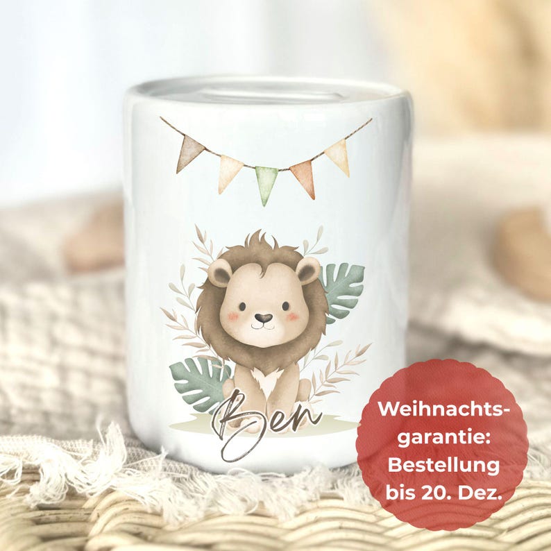 May include: White ceramic money box decorated with a cartoon lion illustration. The lion has a brown mane and sits amongst green leaves. A banner of small flags hangs above the lion. The name "Ben" is written below.