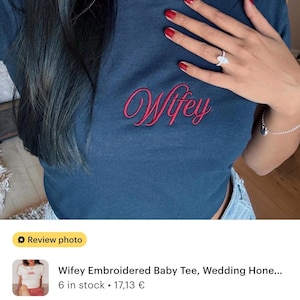 May include: A dark blue short-sleeved crew neck t-shirt featuring the word Wifey embroidered in a red cursive script font across the center of the chest. The garment has a fitted silhouette and a standard ribbed neckline.