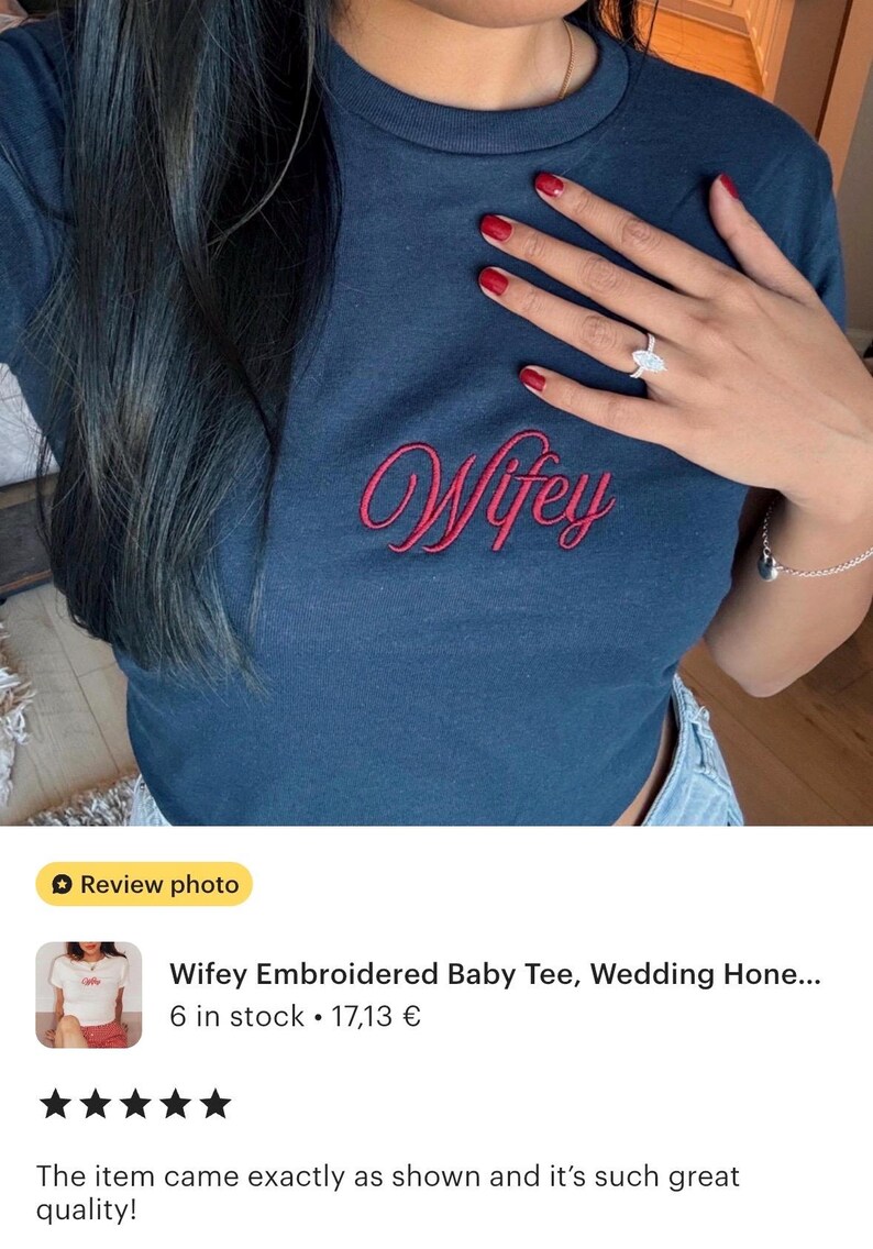 May include: A dark blue short-sleeved crew neck t-shirt featuring the word Wifey embroidered in a red cursive script font across the center of the chest. The garment has a fitted silhouette and a standard ribbed neckline.