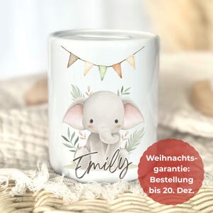 May include: White ceramic money box with a watercolour illustration of a baby elephant, large ears, and the name "Emily". The design includes a banner of small flags and greenery.