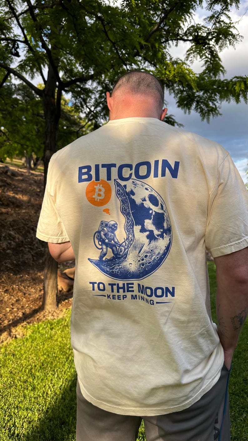 May include: Cream-colored t-shirt with a blue and orange graphic featuring the word "BITCOIN" and an astronaut on the moon. The phrase "TO THE MOON - KEEP MINING" is printed below the graphic.