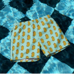 May include: Light blue swim trunks with a repeating pattern of gold Bitcoin symbols. The shorts are made of a lightweight material and are shown against a background of blue water with a black and white checkered pattern.