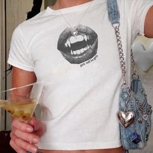 May include: A white short-sleeved t-shirt featuring a black graphic of lips with fangs and the text "girls will be girls" printed below. A silver heart-shaped pendant necklace is worn, and a blue denim shoulder bag with a silver chain strap is present.