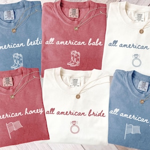 May include: Six t-shirts in shades of blue, red, and white, each with the words "all american" and a different word. Each shirt has a graphic and a gold necklace. The bottom left shirt is crimson.