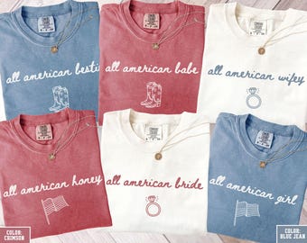 Könnte beinhalten: Sechs T-Shirts in verschiedenen Farben, darunter Blau, Rot und Weiß, mit dem Schriftzug "all american" und verschiedenen Wörtern. Jedes Shirt hat eine Grafik und eine goldene Halskette. Das untere linke Shirt ist karmesinrot.