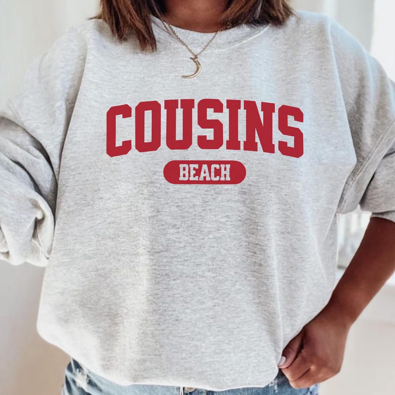 May include: A pale grey sweatshirt featuring the text "COUSINS BEACH" in red, bold lettering. The word "BEACH" is in a red rectangle below "COUSINS". The sweatshirt is a casual style.