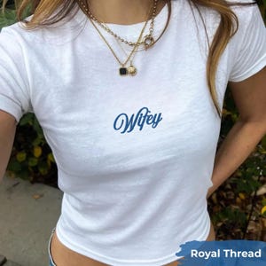 May include: A white short-sleeved t-shirt featuring the word Wifey printed in a blue cursive script across the center of the chest. The shirt is paired with layered gold necklaces, including a chain with a small shell and a black square pendant.