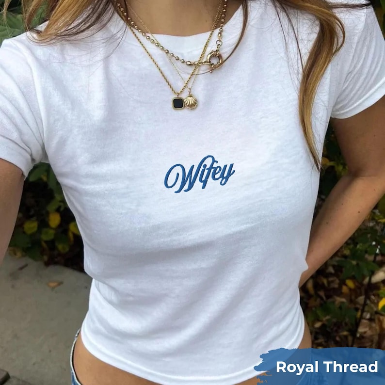 May include: A white short-sleeved t-shirt featuring the word Wifey printed in a blue cursive script across the center of the chest. The shirt is paired with layered gold necklaces, including a chain with a small shell and a black square pendant.