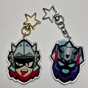 Puede incluir: Dos llaveros acrílicos con diseños de personajes de dibujos animados. Un llavero tiene un cierre dorado en forma de estrella, el otro tiene un cierre plateado en forma de estrella. Los personajes tienen cascos, máscaras y detalles en rojo, negro y azul.