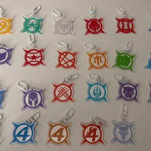 May include: A collection of acrylic keychains featuring various superhero symbols in vibrant colors. Each keychain has a silver clasp for easy attachment. The designs include a hammer, star, and other iconic emblems.