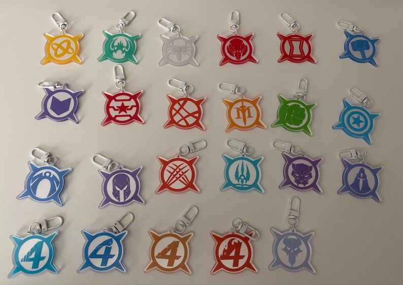 May include: A collection of acrylic keychains featuring various superhero symbols in vibrant colors. Each keychain has a silver clasp for easy attachment. The designs include a hammer, star, and other iconic emblems.