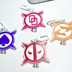 May include: Four acrylic keychains with silver clasps. Each keychain features a different design: a pink "DD" logo, a purple spade, an orange hand design, and a red and white face design. Each keychain has a number printed on it.