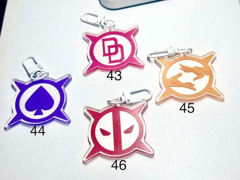 May include: Four acrylic keychains with silver clasps. Each keychain features a different design: a pink "DD" logo, a purple spade, an orange hand design, and a red and white face design. Each keychain has a number printed on it.