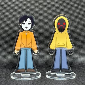 May include: Two acrylic figurines of cartoon characters. One figure wears an orange jacket, blue pants, and a white mask. The other figure is in a yellow hoodie with a black mask and red frown. Both stand on clear circular bases.