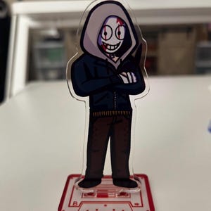 May include: A clear acrylic figure of a character in a dark blue hooded jacket, brown pants, and black shoes. The character has a white mask with a red slash and a wide grin. The figure stands on a red cassette tape base.