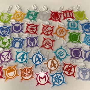 May include: A collection of 30 colourful keychains featuring various superhero logos and symbols. The keychains are made of clear acrylic with a starburst design around each logo. The logos include characters such as Iron Man, Spider-Man, Captain America, and more.