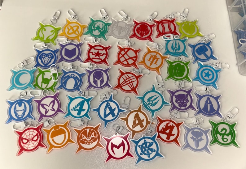 May include: A collection of colorful acrylic keychains featuring various superhero symbols. Each keychain has a silver clasp and a unique design in shades of red, blue, green, orange, and purple. The keychains are arranged on a white surface.
