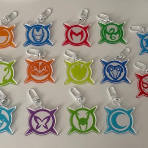 May include: A collection of colorful acrylic keychains with various superhero-themed designs. Each keychain features a unique emblem in orange, blue, red, green, and purple, attached to a silver clasp. The designs include a shark, butterfly, and spider.