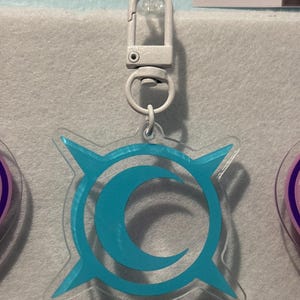 May include: A clear acrylic keychain with a light blue crescent moon and starburst design. The keychain has a silver-colored clasp for easy attachment. The design is centered on the keychain, and the overall aesthetic is modern and minimalist.